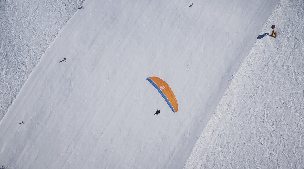 Paraglider above ski slope | © Davos Klosters Mountains An orange paraglider flies over a ski slope that lies in a straight line below it. | © Davos Klosters Mountains