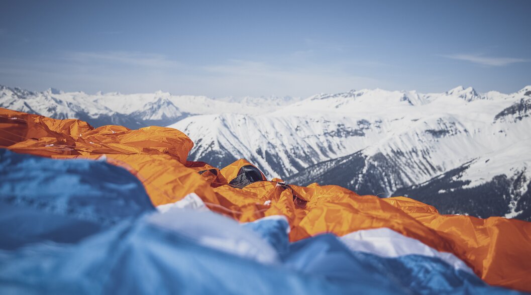 Close-up paraglider | © Davos Klosters Mountains Close-up of a blue-orange paraglider laid out on the ground with snow-covered mountains in the background. | © Davos Klosters Mountains