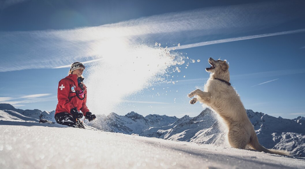 Seraina und Lawinenhünding Emma im Schnee | © Davos Klosters Mountains Patrouilleurin Seraina wirft ihrer Golden Retriever Hündin Emma Schnee in die Luft. | © Davos Klosters Mountains