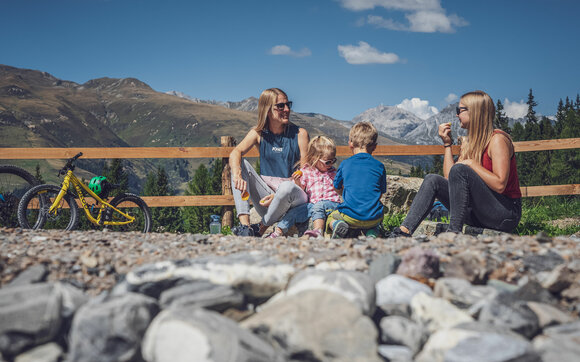 Zwei Frauen und zwei Kinder sitzen am See auf dem Rinerhorn und stärken sich mit einem Snack. | © Davos Klosters Mountains