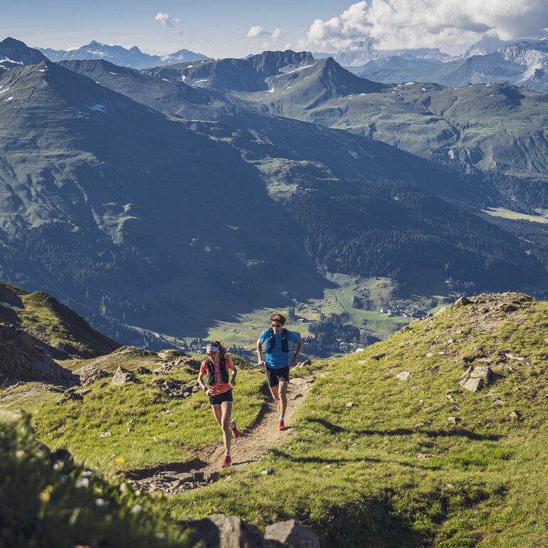 A trailrunning couple is running on the ridge of the mountain Jakobshorn, down in the valley the lake of Davos is apparent.  | © Davos Klosters Mountains