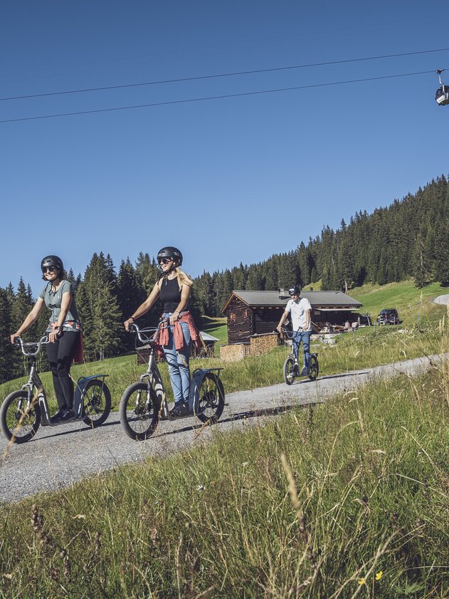 Scooter downhill Rinerhorn gondolas | © Davos Klosters Mountains A group of friends ride their scooters under the Rinerhorn gondola, past a green meadow towards the valley. | © Davos Klosters Mountains