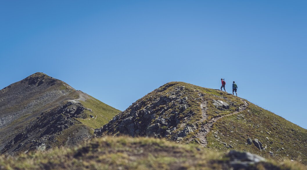 In der Ferne wandert ein Paar auf dem Grat des Jakobshorns in Richtung des Jatzhorns und geniesst den wolkenlosen Himmel.  | © Davos Klosters Mountains