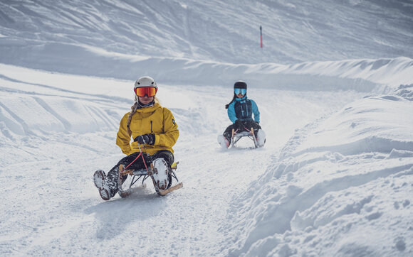 Zwei Freundinnen in farbiger Winterkleidung schlitteln auf der Schlittelstrecke Rinerhorn ins Tal. | © Davos Klosters Mountains