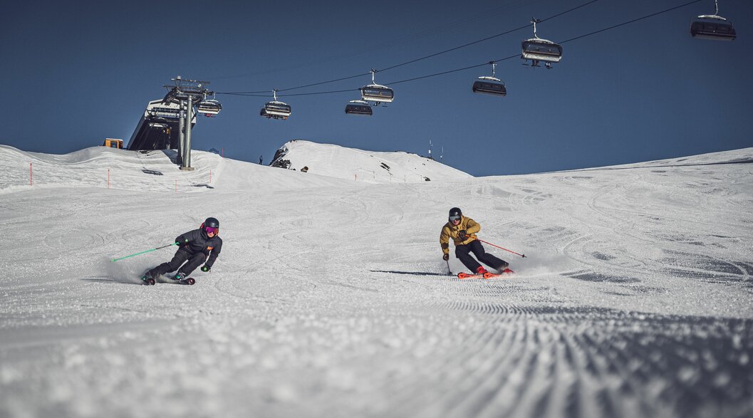 Eine Skifahrerin und ein Skifahrer fahren über die Skipiste auf Parsenn, der Sessellift Rapid im Hintergrund. | © Davos Klosters Mountains